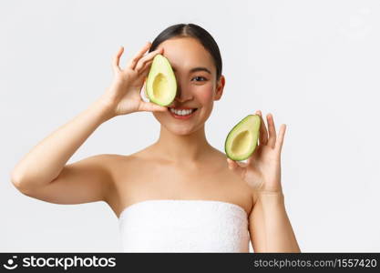 Beauty, personal care, spa and skincare concept. Close-up of tender feminine asian woman in bath towel, smiling and showing avocado near face, advertisement of face mask, cleanser or cream.. Beauty, personal care, spa and skincare concept. Close-up of tender feminine asian woman in bath towel, smiling and showing avocado near face, advertisement of face mask, cleanser or cream