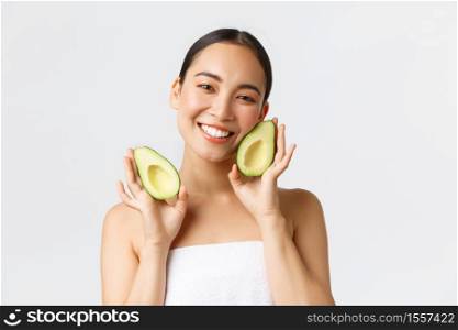 Beauty, personal care, spa and skincare concept. Tender cute asian female in bath towel showing avocado and smiling satisfied, nourish and treat skin with face masks or cream, white background.. Beauty, personal care, spa and skincare concept. Tender cute asian female in bath towel showing avocado and smiling satisfied, nourish and treat skin with face masks or cream, white background