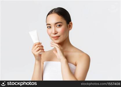 Beauty, personal care, spa salon and skincare concept. Young beautiful asian woman in bath towel touching face and showing facial cream, moisturizing treatment of skin, standing white background.. Beauty, personal care, spa salon and skincare concept. Young beautiful asian woman in bath towel touching face and showing facial cream, moisturizing treatment of skin, standing white background