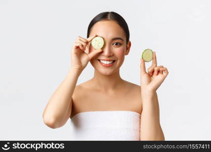 Beauty, personal care, spa salon and skincare concept. Beautiful young asian female in bath towel holding cucumbers and smiling, promo of facial or body treatment, moisturizing features of cream.. Beauty, personal care, spa salon and skincare concept. Beautiful young asian female in bath towel holding cucumbers and smiling, promo of facial or body treatment, moisturizing features of cream
