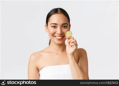Beauty, personal care, spa salon and skincare concept. Pretty asian female in bath towel smiling and holding cucumber piece, promo of cleansing gel, face mask, moisturizer or body gel for shower.. Beauty, personal care, spa salon and skincare concept. Pretty asian female in bath towel smiling and holding cucumber piece, promo of cleansing gel, face mask, moisturizer or body gel for shower