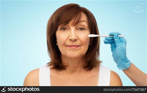 beauty, plastic surgery and cosmetology concept - senior woman and surgeon hands with scalpel over blue background. senior woman and surgeon hands with scalpel