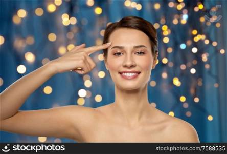beauty, plastic surgery and people concept - beautiful young woman with bare shoulders showing her forehead over holidays lights on dark blue background. beautiful young woman showing her forehead