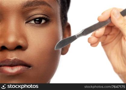 beauty, plastic surgery and people concept - close up of face of beautiful young african american woman and hand with scalpel knife over white background. face of african american woman and scalpel knife