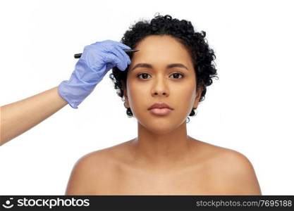 beauty, plastic surgery and people concept - close up of face of beautiful young african american woman and hand in glove with marker over white background. face of african woman and hand with marker