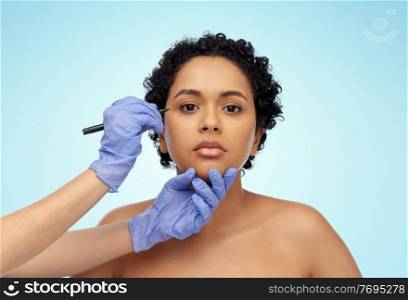 beauty, plastic surgery and people concept - close up of face of beautiful young african american woman and hands in gloves with marker over blue background. face of african woman and hands with marker