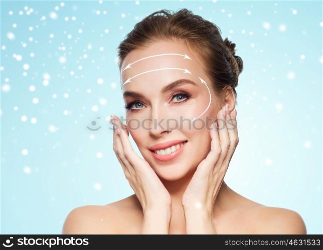 beauty, plastic surgery, facelift, people and rejuvenation concept - beautiful young woman touching her face with lifting arrows over blue background and snow