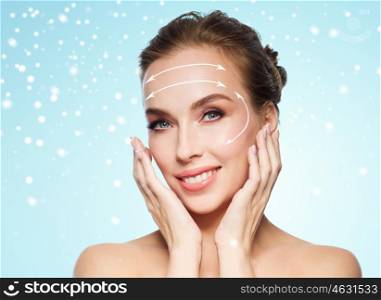 beauty, plastic surgery, facelift, people and rejuvenation concept - beautiful young woman touching her face with lifting arrows over blue background and snow