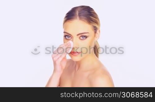 Beauty portrait of a gorgeous young woman with blue eyes cleansing her face with a cotton pad looking at the camera with a beautiful smile
