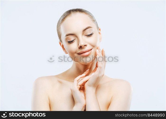 Beauty portrait of attractive topless woman touches cheek gently, enjoys softness of skin, has natural makeup, manicure, cares about body and complexion, isolated on white background. Wellness concept