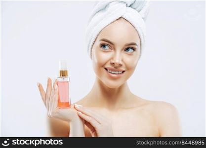 Beauty portrait of happy smiling European woman with healthy skin, makeup, holds bottle of aromatic parfume, stands naked indoor, wears towel on head. Women, cosmetology and skin care concept