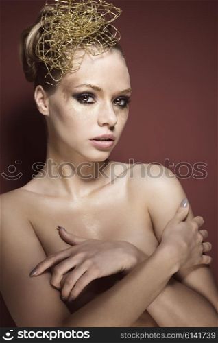 beauty portrait of sexy blonde woman with glossy creative golden make-up and accessory in the hair-style. She is posing with nude shoulders, covering her breast and looking in camera with sensual eyes