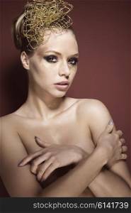 beauty portrait of sexy blonde woman with glossy creative golden make-up and accessory in the hair-style. She is posing with nude shoulders, covering her breast and looking in camera with sensual eyes