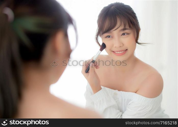 Beauty portrait young asian woman smile with face looking mirror applying makeup with brush cheek in the bedroom, beautiful of girl holding blusher, skin care and cosmetic concept.