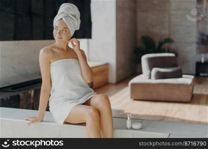 Beauty procedure, skin care and home treatment concept. Dreamy satisfied young woman with healthy skin, enjoys smoothing skin, domestic routine, uses cosmetic products, wrapped in bath towel