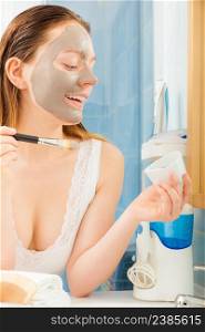 Beauty procedures skin care concept. Young woman applying facial gray mud clay mask to her face in bathroom