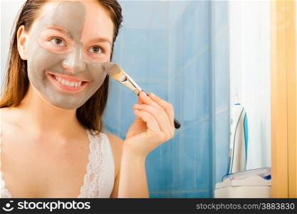 Beauty procedures skin care concept. Young woman applying facial gray mud clay mask to her face in bathroom