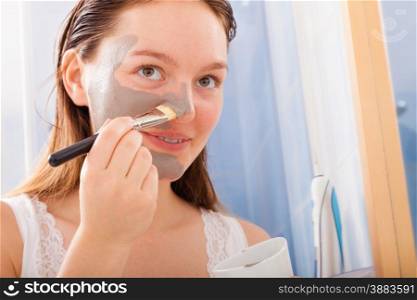 Beauty procedures skin care concept. Young woman no makeup applying facial gray mud clay mask to her face in bathroom using brush