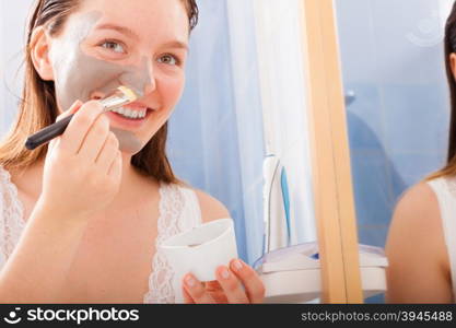 Beauty procedures skin care concept. Young woman no makeup applying facial gray mud clay mask to her face in bathroom using brush