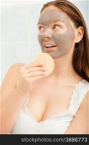Beauty procedures spa and skin care concept. Young woman with facial clay mask in bathroom holds sponge to remove mud