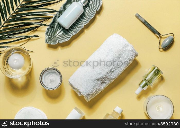 Beauty products for skin care on yellow background with palm leaf.  Facial cosmetics with towel, jade massage roller, candles, pipette bottles with serum and essential oil. Top view.