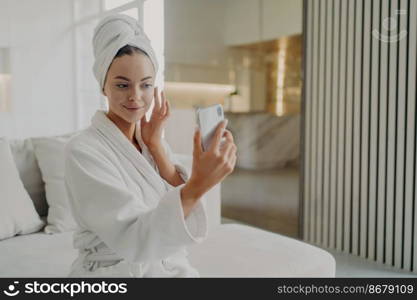 Beauty routine. Young gorgeous woman touching her soft healthy skin, applying cosmetic cream while sitting on sofa, female wearing bathrobe and towel on head holding smartphone in front of her face. Young beautiful woman applying cosmetic cream while relaxing on sofa after taking shower at home