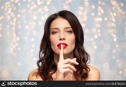beauty, secret, make up and people concept - woman holding finger on lips or mouth with red lipstick over holidays lights background. woman with red lipstick holding finger on mouth. woman with red lipstick holding finger on mouth