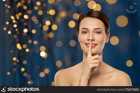 beauty secret, silence and people concept - beautiful young woman holding finger on lips over holidays lights on dark blue background. beautiful young woman holding finger on lips