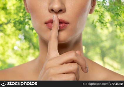beauty secret, silence and people concept - close up of beautiful young woman holding finger on lips over green natural background. beautiful young woman holding finger on lips