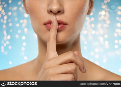 beauty secret, silence and people concept - close up of beautiful young woman holding finger on lips over holidays lights on blue background. beautiful young woman holding finger on lips