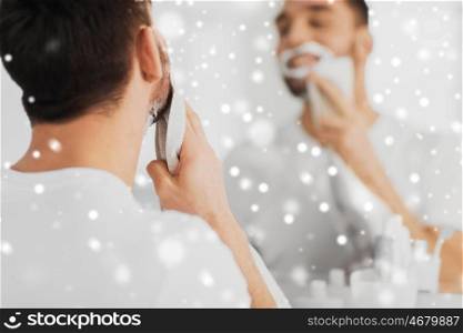 beauty, shaving, grooming and people concept - close up of man removing shaving foam from face with towel and looking to mirror at home bathroom over snow