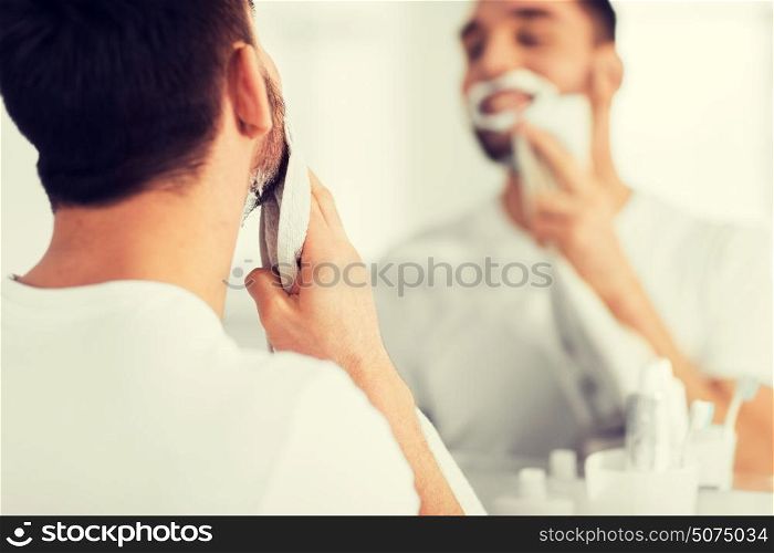 beauty, shaving, grooming and people concept - close up of man removing shaving foam from face with towel and looking to mirror at home bathroom. close up of man removing shaving foam from face