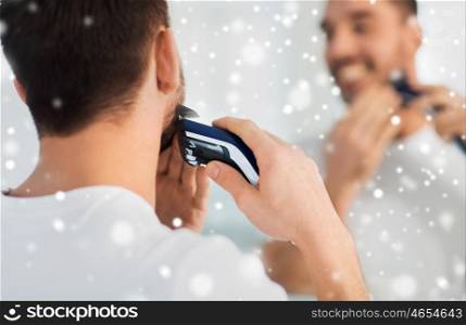 beauty, shaving, grooming and people concept - close up of young man looking to mirror and shaving beard with trimmer or electric shaver at home bathroom over snow