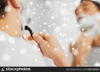 beauty, shaving, grooming and people concept - close up of young man looking to mirror and shaving beard with manual razor blade at home bathroom over snow