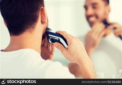 beauty, shaving, grooming and people concept - close up of young man looking to mirror and shaving beard with trimmer or electric shaver at home bathroom. close up of man shaving beard with trimmer