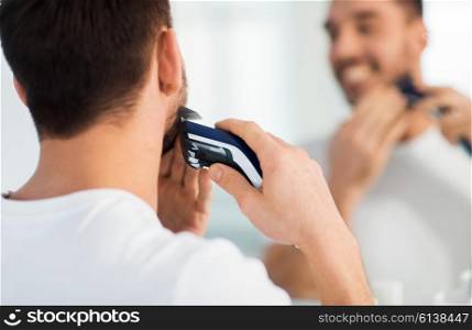 beauty, shaving, grooming and people concept - close up of young man looking to mirror and shaving beard with trimmer or electric shaver at home bathroom
