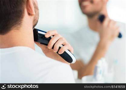 beauty, shaving, grooming and people concept - close up of young man looking to mirror and shaving beard with trimmer or electric shaver at home bathroom