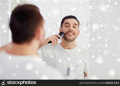 beauty, shaving, grooming and people concept - young man looking to mirror and shaving beard with trimmer or electric shaver at home bathroom over snow