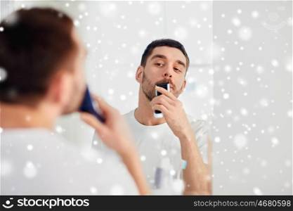 beauty, shaving, grooming and people concept - young man looking to mirror and shaving beard and mustache with trimmer or electric shaver at home bathroom over snow