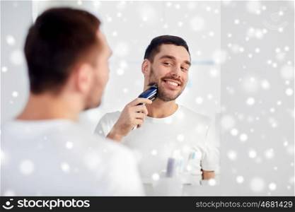 beauty, shaving, grooming and people concept - young man looking to mirror and shaving beard with trimmer or electric shaver at home bathroom over snow