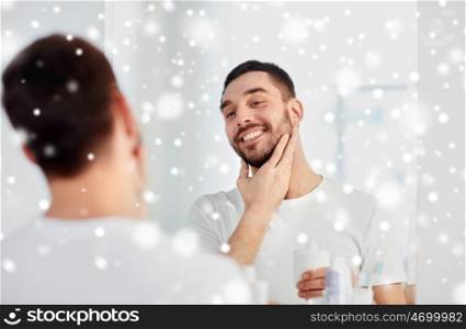 beauty, shaving, grooming, winter and people concept - smiling young man looking to mirror and applying aftershave at home bathroom over snow