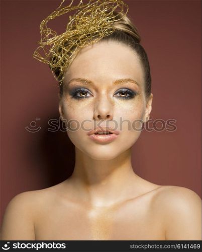 beauty shoot of very pretty woman with glossy golden creative make-up and accessory in the hairdo. She is covering her nude breast and looking in camera