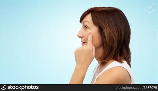 beauty, skin aging and old people concept - profile of senior woman pointing to her eye wrinkles over blue background. profile of senior woman pointing to eye wrinkles