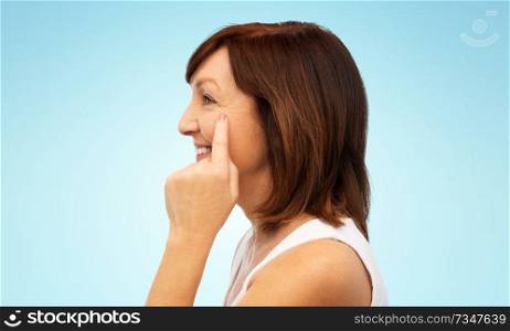 beauty, skin aging and old people concept - profile of smiling senior woman pointing to her eye wrinkles blue white background. profile of senior woman pointing to eye wrinkles