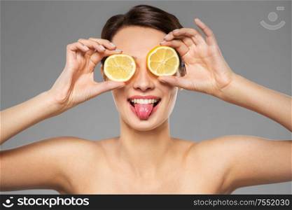 beauty, skin care and detox concept - beautiful woman making eye mask of lemon slices and showing her tongue over grey background. beautiful woman making eye mask of lemon slices