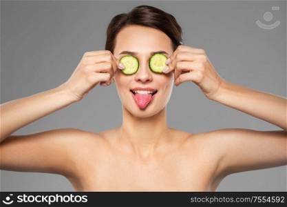 beauty, skin care and detox concept - beautiful woman making eye mask of cucumbers and showing her tongue over grey background. beautiful woman making eye mask of cucumbers