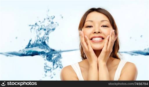 beauty, skin care and hydration concept - happy smiling young asian woman touching her face over blue background with water splash. happy smiling young asian woman touching her face