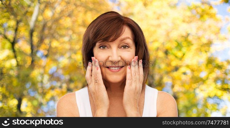 beauty, skin care and old people concept - portrait of smiling senior woman touching her face over natural autumn background. smiling senior woman touching her face in autumn
