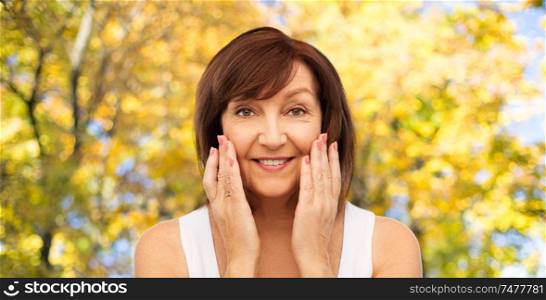 beauty, skin care and old people concept - portrait of smiling senior woman touching her face over natural autumn background. smiling senior woman touching her face in autumn