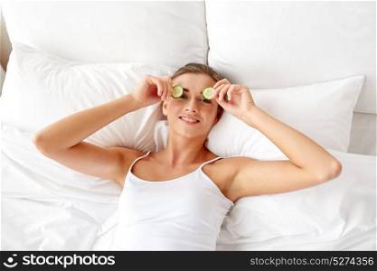beauty, skin care and people concept - beautiful young woman lying in bed and applying cucumbers to her eyes at home bedroom. beautiful woman applying cucumbers to eyes at home